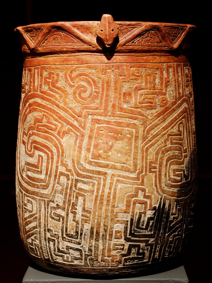 Ceramic cylinder-shaped vessel decorated with geometric carvings and face at the rim.