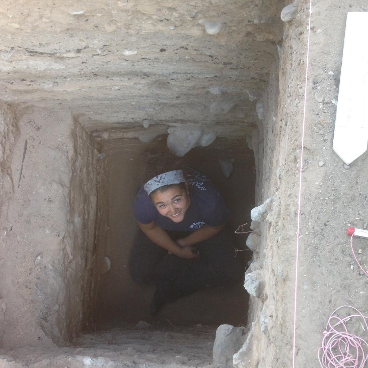 Person sitting in deep, square hole while looking up at camera, smiling.