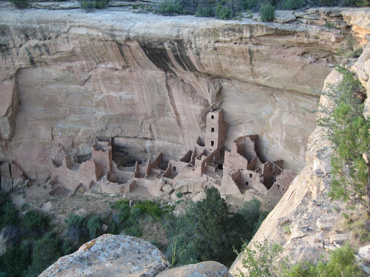 Round and square interconnected stone buildings built against a cliff face. One is 4 stories tall.