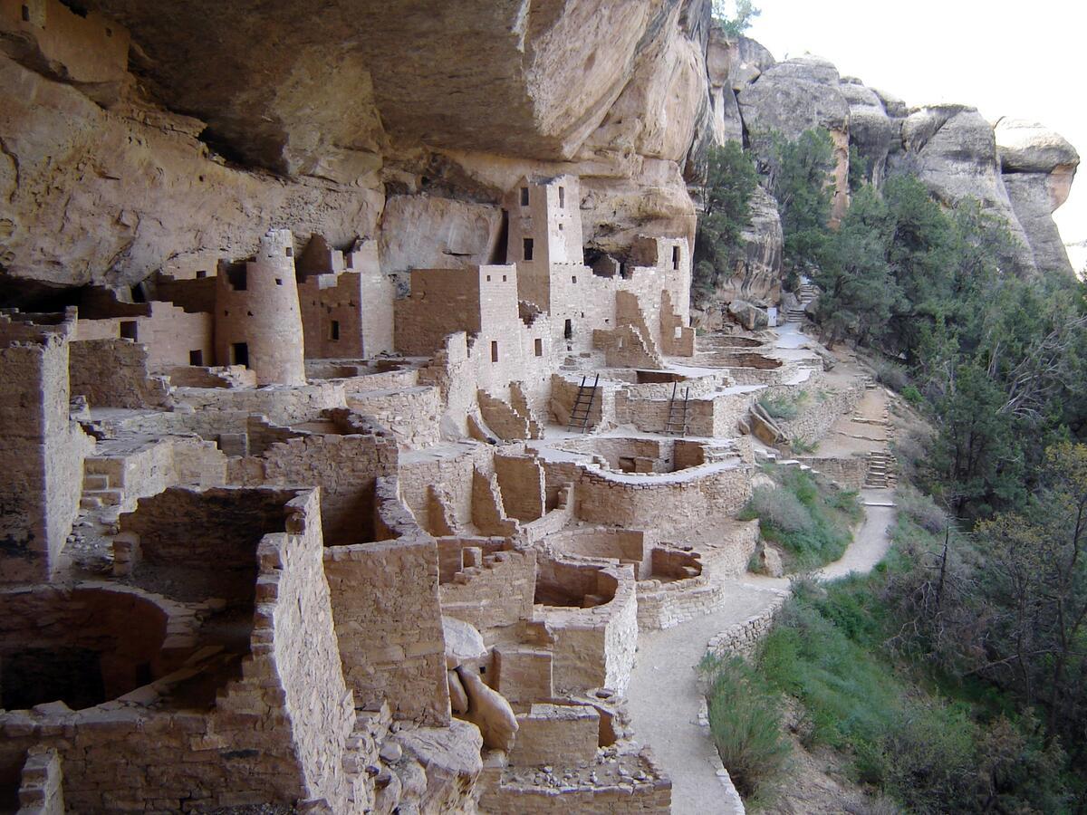 Multi-story stone buildings built under a cliff overhang on the edge of a steep cliff.